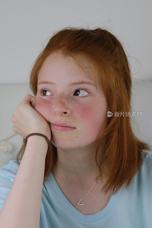 这是一个14 / 15岁的红发少女的照片，她皮肤苍白，脸上有雀斑，脸颊通红，坐在卧室里，看起来厌倦了，下巴靠在手上，看起来闷闷不乐